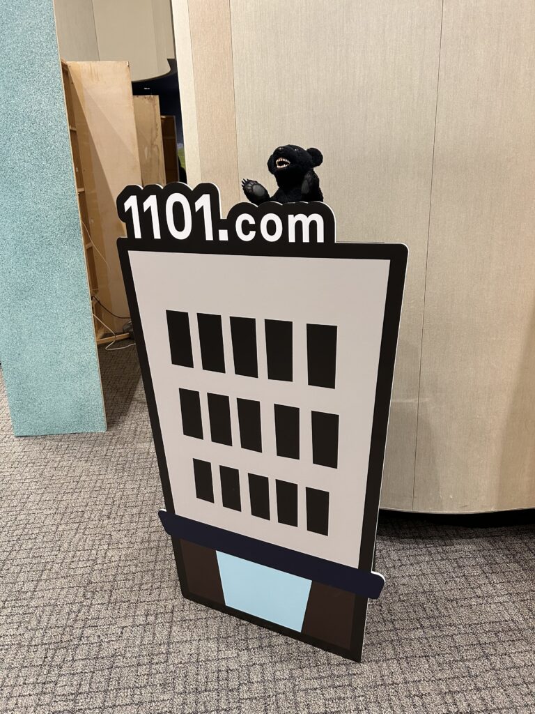 ほぼ日の株主総会2025で展示されていた1101.comのビルのイラストパネルとクマのぬいぐるみ
